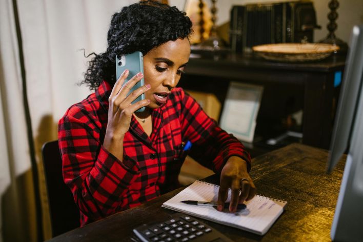 Focused woman multitasks at her desk, talking on the phone and writing notes.