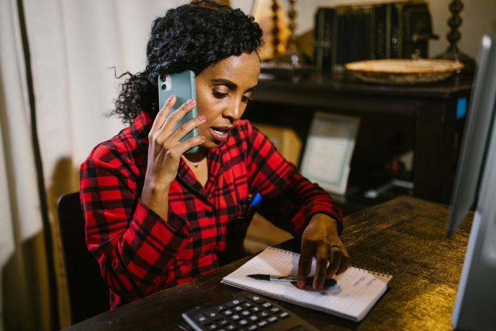 Focused woman multitasks at her desk, talking on the phone and writing notes.