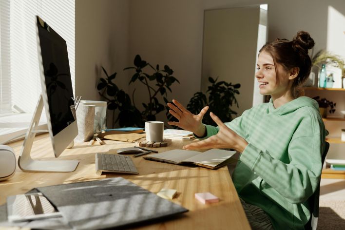A woman in a green hoodie having a video call at her home office desk.